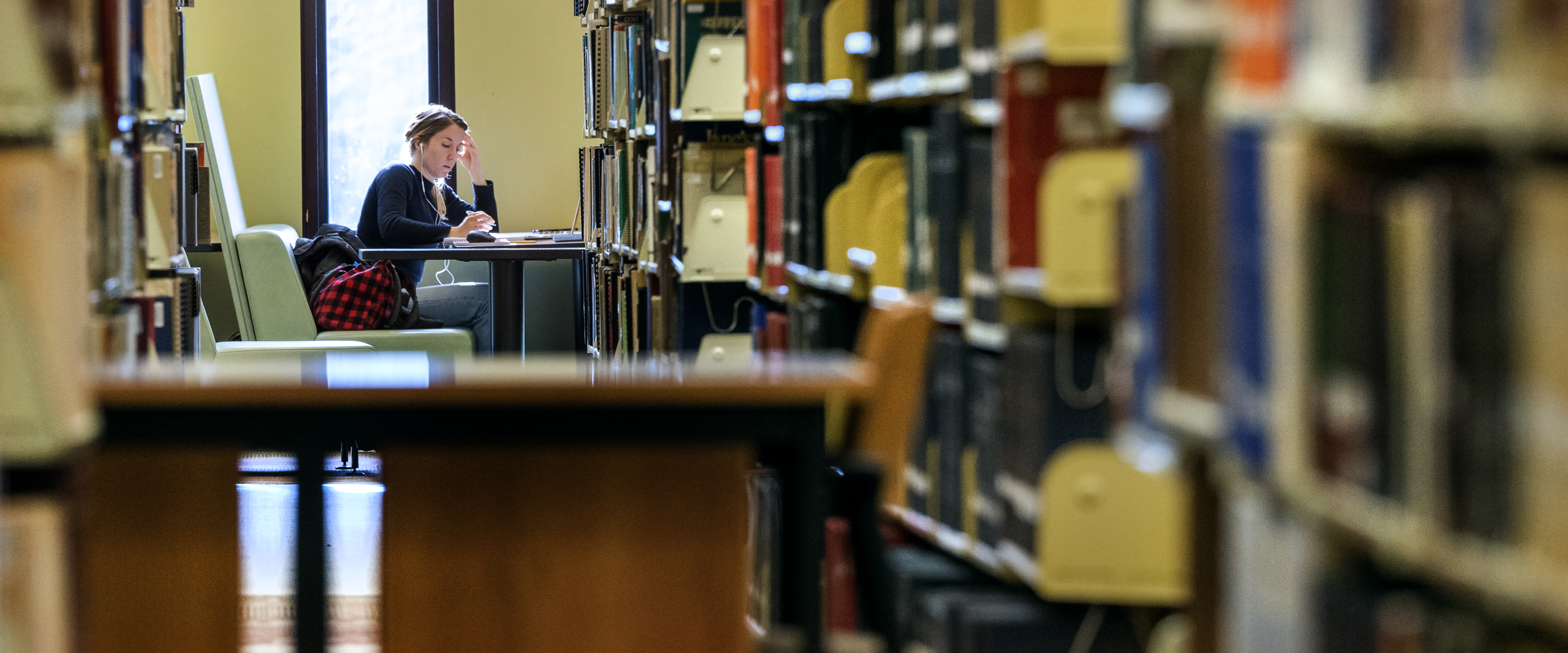 A student sits a table, near a window in a library, studying.