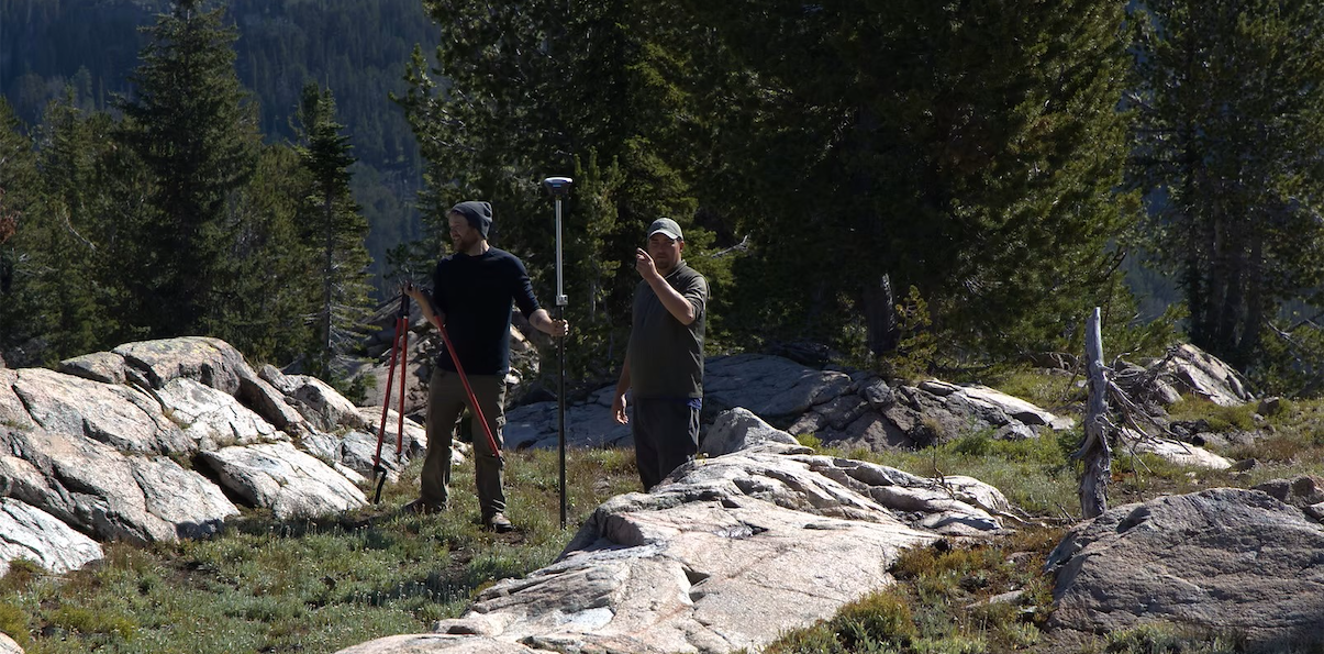 Two people stand near rocks and trees; one person holds tools while the other person points to something out of frame.