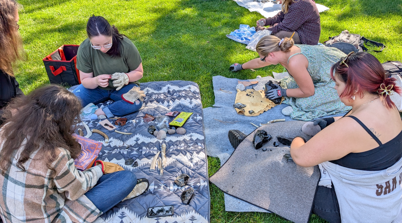Students sit on a blanket outdoors wearing gloves and attempting to break pieces of obsidian into sharp tools.