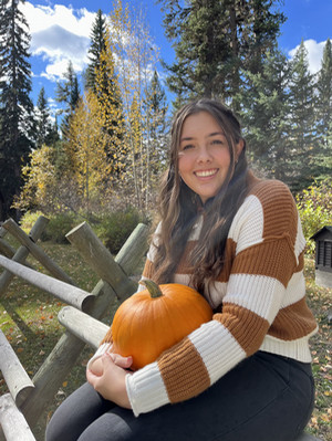 ASUM Business Manager Hailey Vondal in a stripped sweater holding a pumpkin and sitting on a jack-leg fence in rural Montana in the fall. 