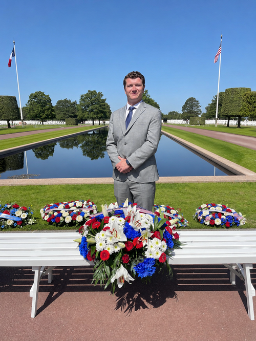 ASUM Vice President Lane Allen standing in a grey suit at the World War two Memorial in Normandy, France with Red, White, and Blue flower wreaths. 