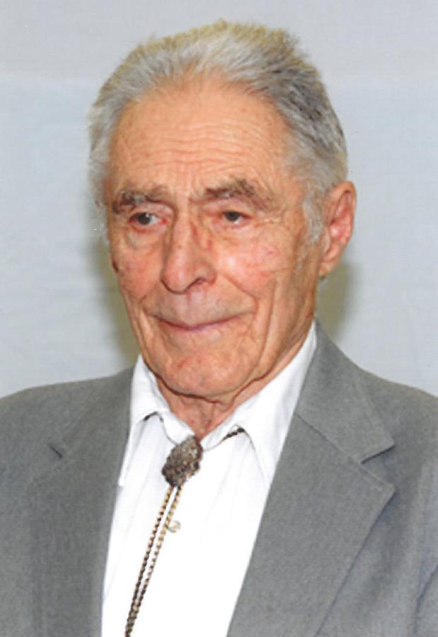 Portrait of Bill Morrelles with gray hair wearing a light gray suit jacket, white shirt, and bolo tie against a plain background.