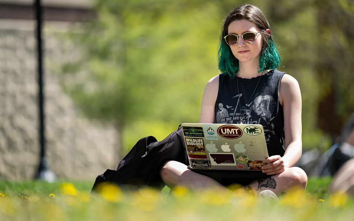 A UM student works on a laptop while sitting in the grass