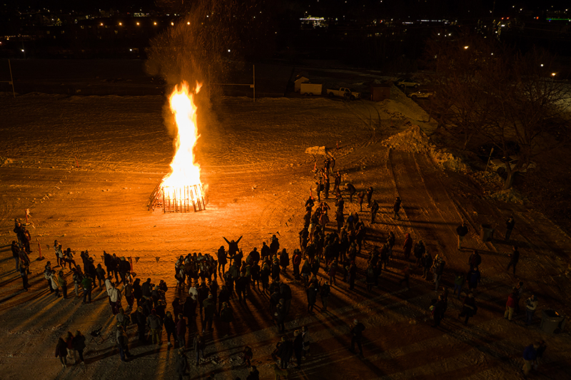 A bonfire rages while a large crowd stands a safe distance away