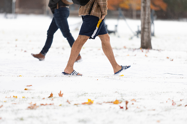 A student wearing shorts and flipflops trudges through the snow while crossing campus.