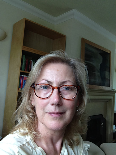 Cynthia Larbey with blonde hair and glasses looking at the camera in a living room with a bookshelf in the background.