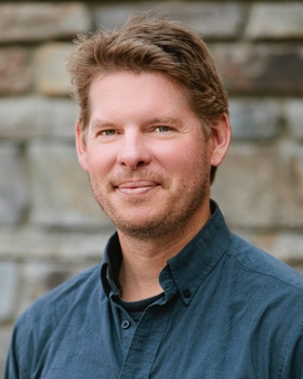 portrait of a smiling Eric Carlson in a dark button-up shirt against a stone wall background.