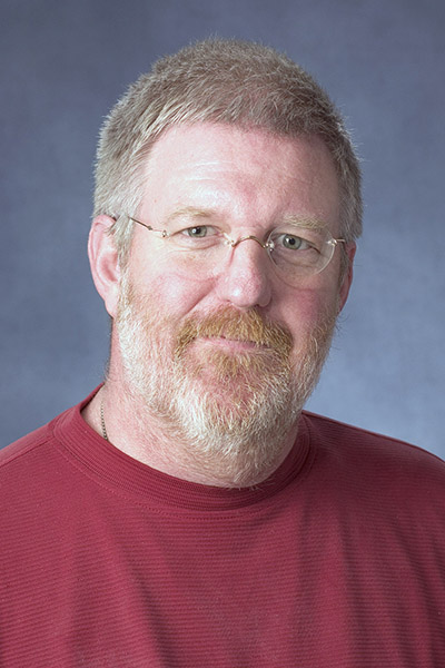 portrait of Dr. Thomas Foor with glasses and a gray beard wearing a red shirt against a blue background.