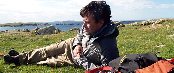 Dr. Nathan Goodale resting on grassy coastal hillside beside a backpack, looking out toward the ocean and distant islands.