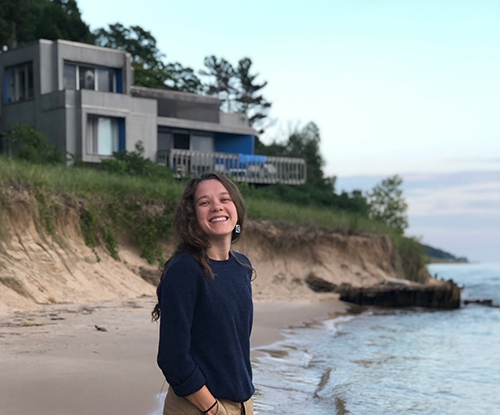 Jean Larmon smiling and standing on a sandy beach near the shoreline, with modern houses on the bluff behind.