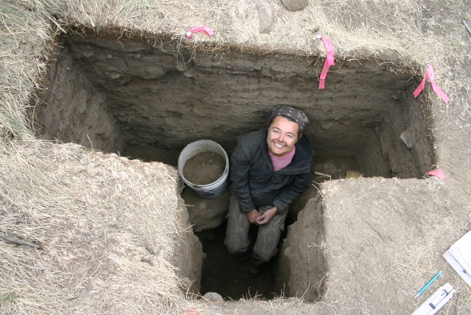 Dr. Lisa Smith smiling while standing in an excavated dirt pit, with a bucket of soil beside them.