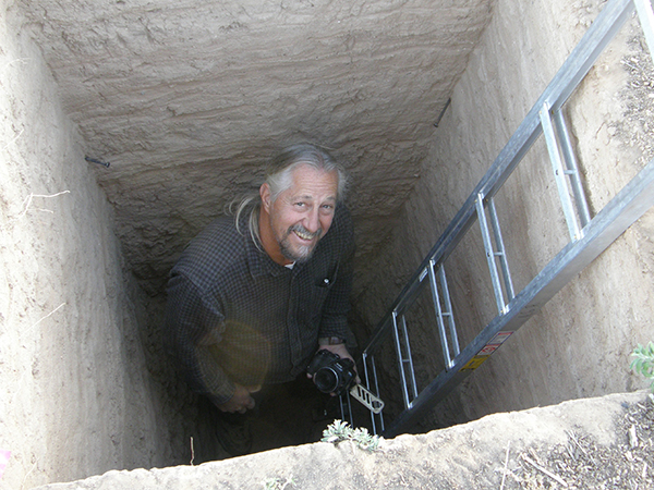 Dr. Matthew Schmader smiling up from inside a deep rectangular pit next to a metal ladder, holding a camera.