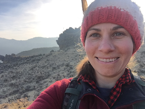 Smiling Nicole Herzog in a knit hat and red jacket taking a selfie on a rocky mountain trail at sunrise.