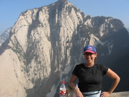 Smiling Dr. Pei-Lin Yu in a cap posing on a ledge with a steep granite mountain in the background.