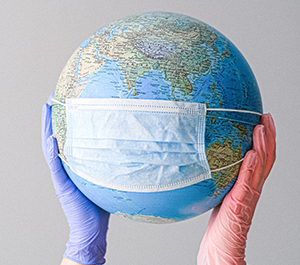 A globe with a medical mask wrapped around it is held up by a person wearing latex gloves