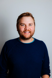 Ryan Hansen smiling with a short beard wearing a navy sweater against a light gray background.