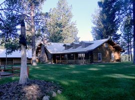 Rustic log cabin lodge with a covered porch, surrounded by tall pine trees and a grassy lawn.