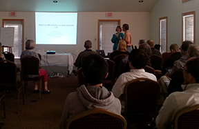 Audience seated in a conference room watching a presentation while two speakers stand near a projected slide.