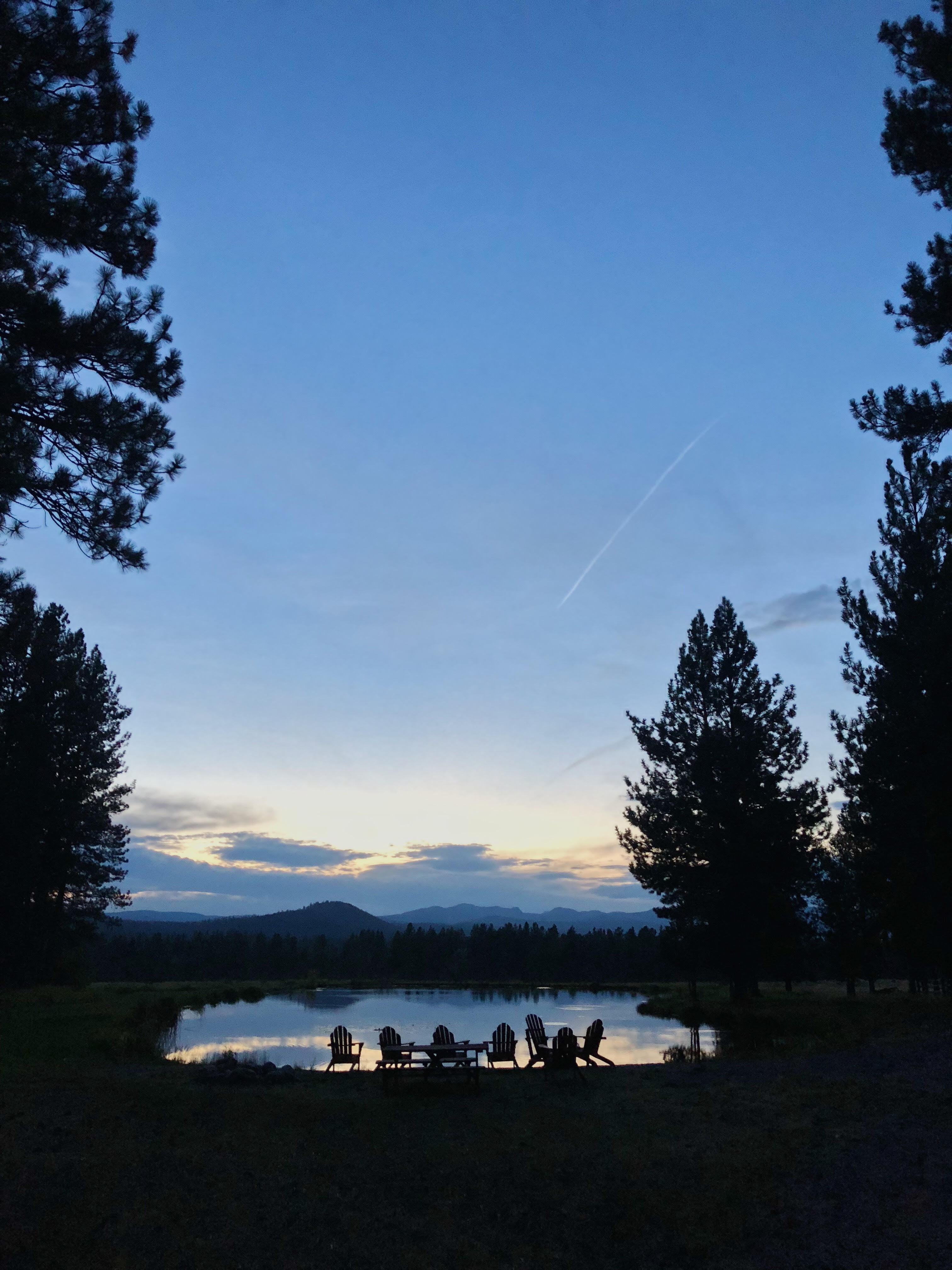Silhouetted Adirondack chairs by a pond at sunset, framed by pine trees with distant mountains on the horizon.