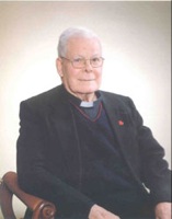 Portrait of Father Robert Zahn in dark clothing seated in a chair against a light background.