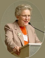 Jean’ne Shreeve speaking at a podium with a microphone, wearing a beige blazer and orange top.