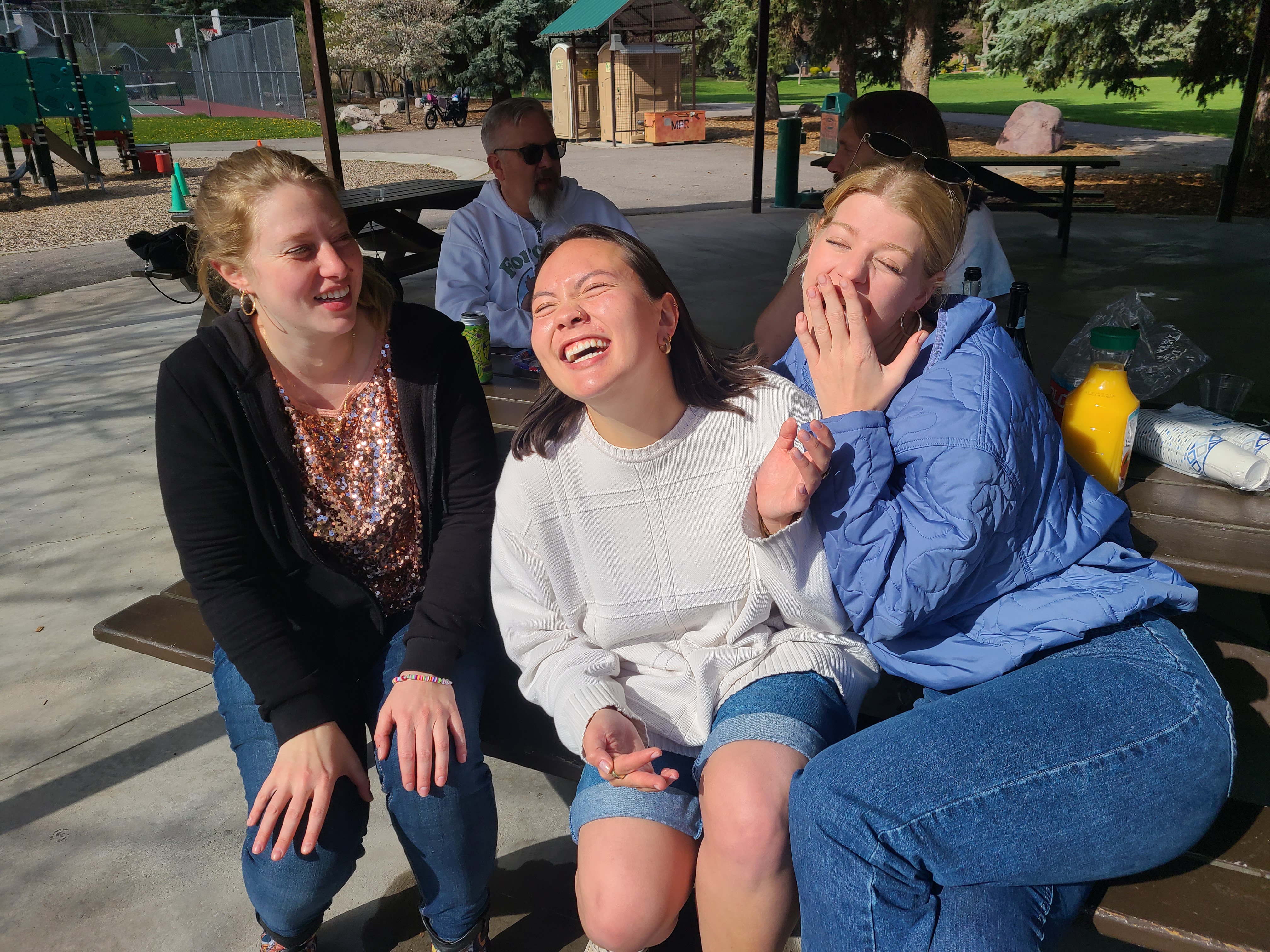 Three students laughing and joking outdoors, sitting at a picnic table. 