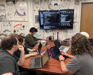 Small group working on laptops around a conference table in a room with a wall of hand-drawn portraits and a large monitor displaying code.