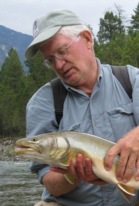 Ted Smith holding a large fish while standing by a river with forested mountains in the background.