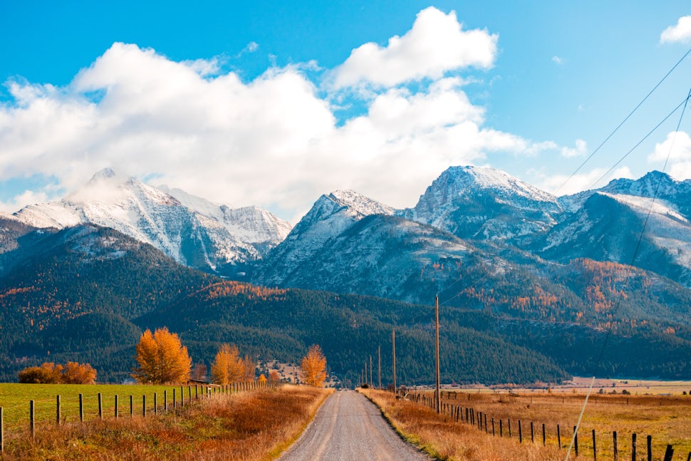 Rural gravel road leading through autumn fields toward snow-capped mountains under a bright blue sky with scattered clouds.