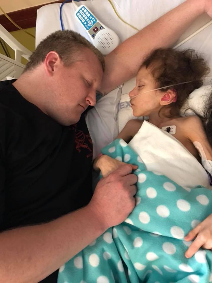 Man sleeping beside a young girl in a hospital bed, holding her hand as she rests with an oxygen tube.