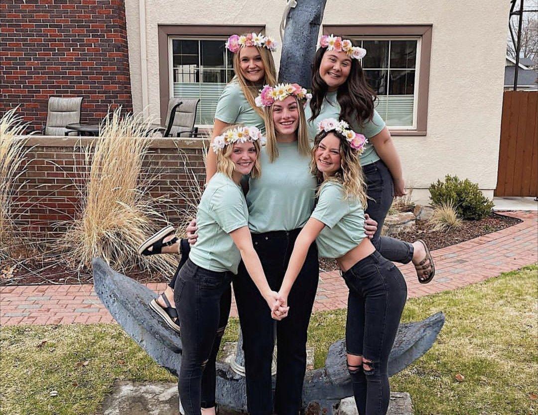 Five sorority members wearing flower crowns pose together outdoors around a large anchor sculpture.