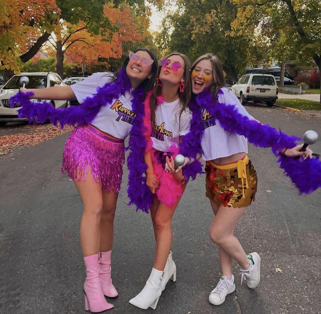 Three members of kappa alpha theta in sparkly skirts and sunglasses pose on a neighborhood street with purple feather boas and fall trees in the background.