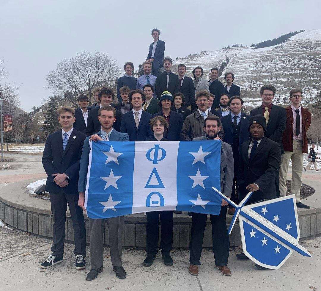 Group of men in suits posing outdoors in winter, holding a blue-and-white Phi Delta Theta flag and shield sign.