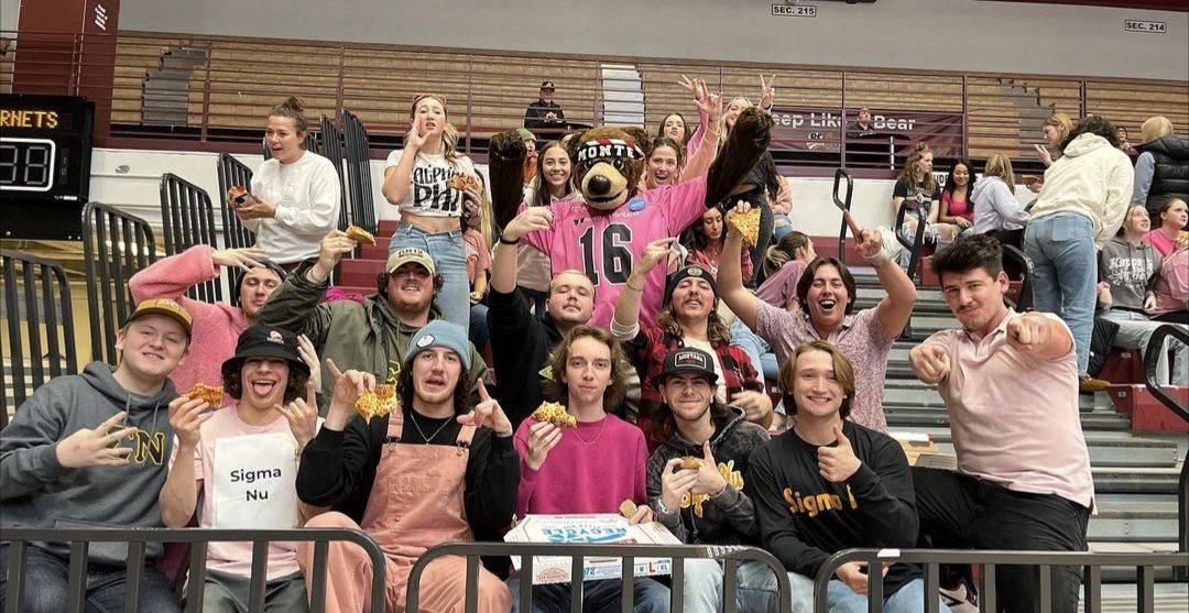 members of sigma nu posing with the mascot Monte on some bleachers with pizza