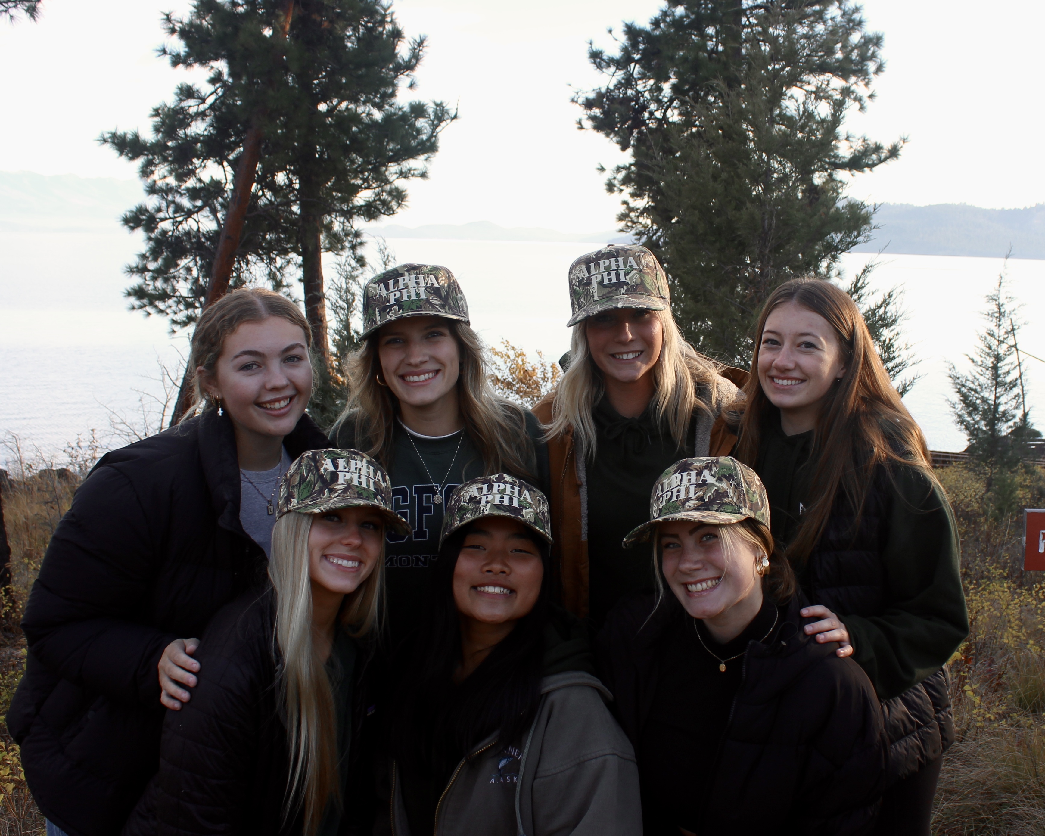 Group of Alpha phi members smiling outdoors by a lakeside viewpoint, some wearing camo Alpha Phi hats.