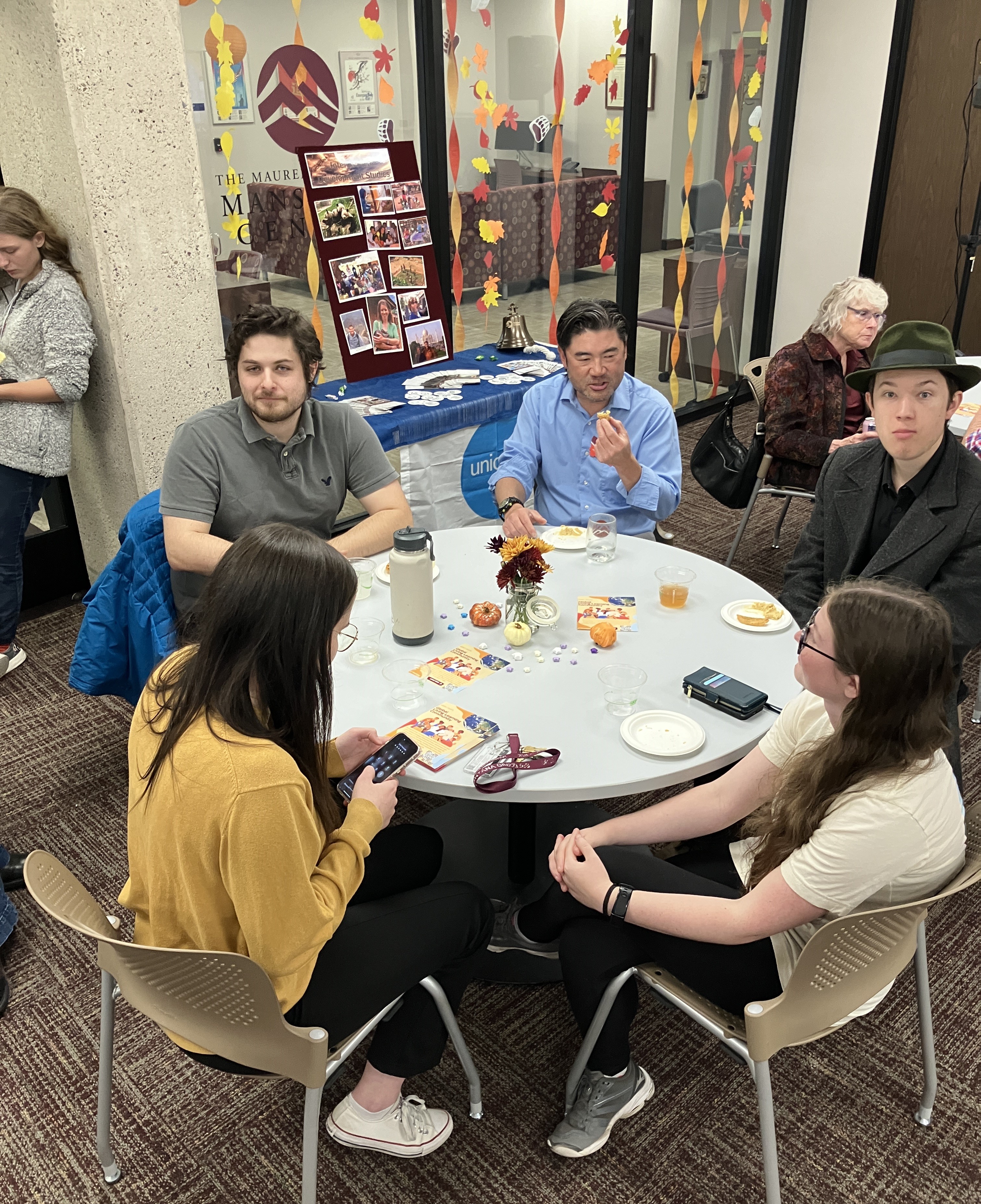Group of staff and students share pleasantries and snacks at a circular table.