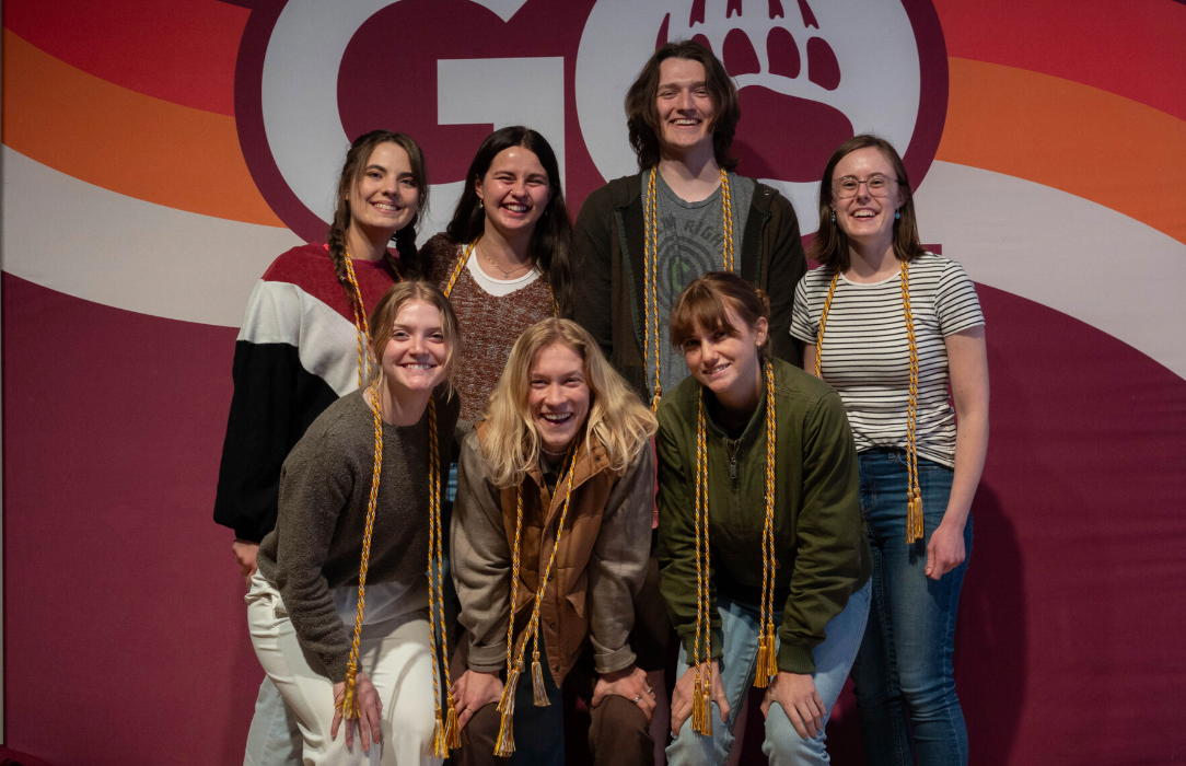 seven university students smiling in front of a go griz sign with their graduation cords