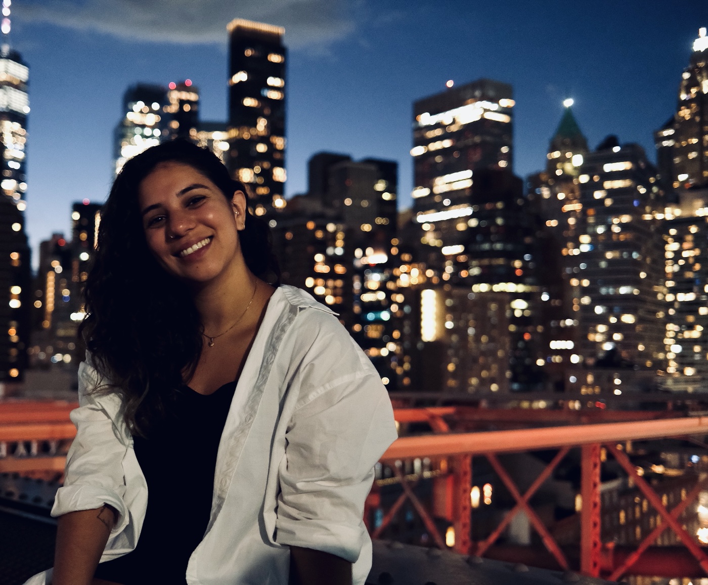 Smiling Ana Carolina posing on a bridge at night with a city skyline of illuminated skyscrapers in the background.