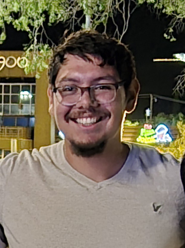 Smiling Arturo Jimenez wearing glasses in a light gray T-shirt outdoors at night with trees and lights in the background.