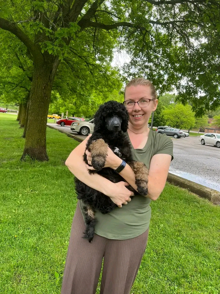 Deborah Ith holding a black-and-tan puppy in a grassy park with trees and parked cars in the background.