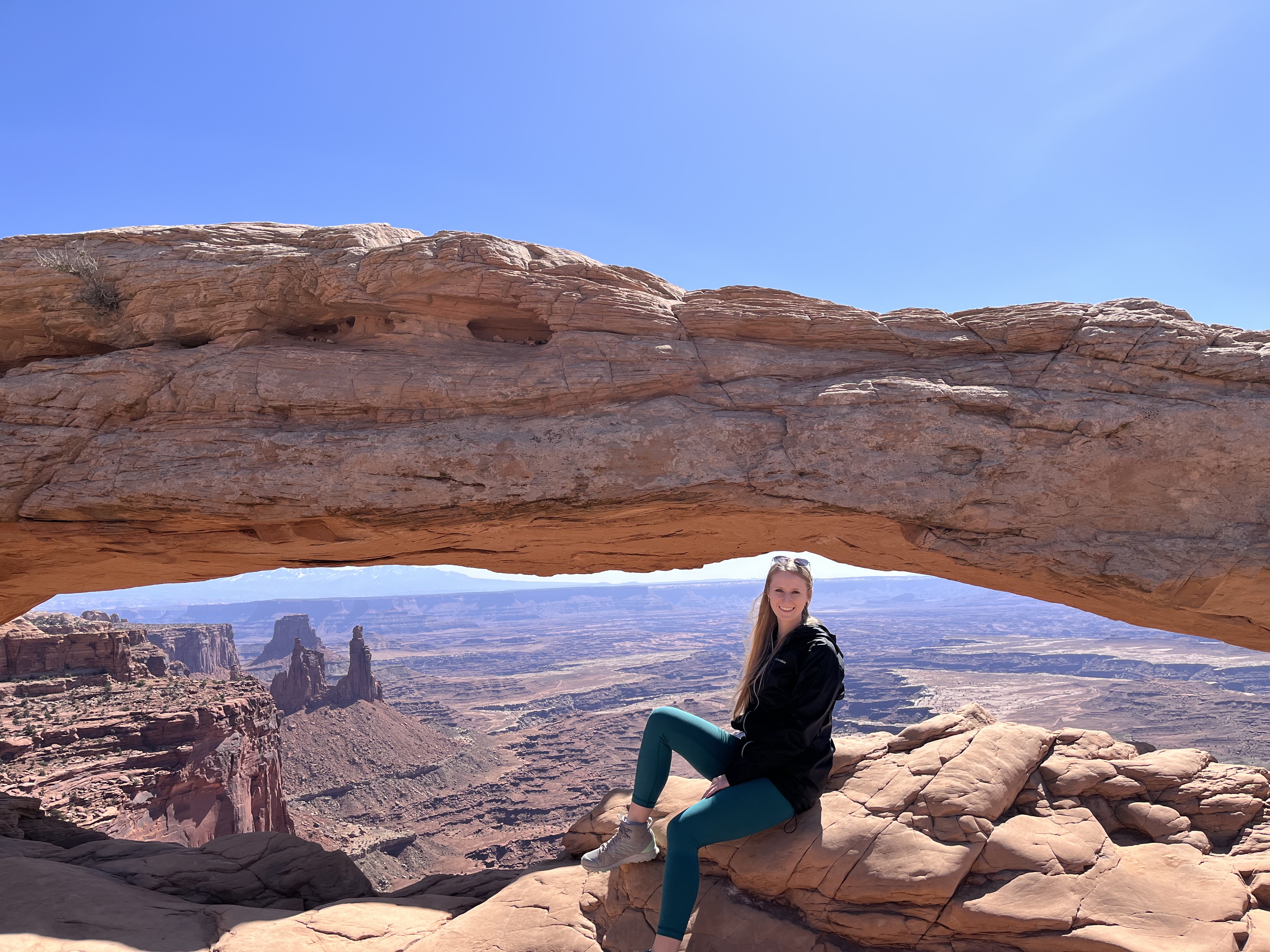 Anna Reszewicz sitting on a rock ledge beneath a large desert sandstone arch, overlooking a canyon landscape.