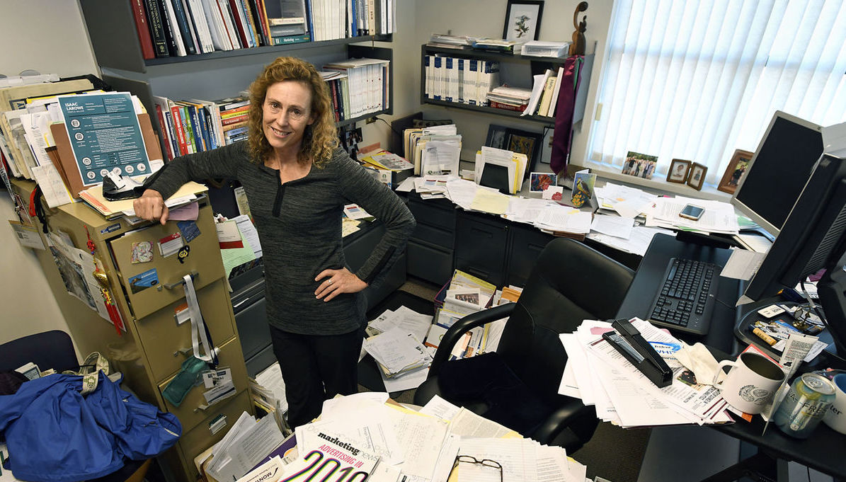 A woman smiles with her arms folded. She stands in a office covered in piles of papers.