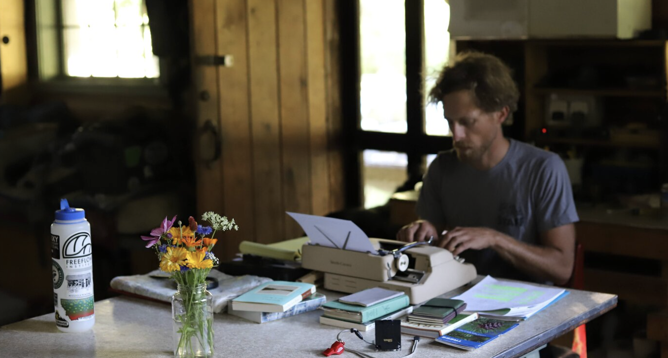 A man types at a typewriter on a desk full of papers, books, and journals.