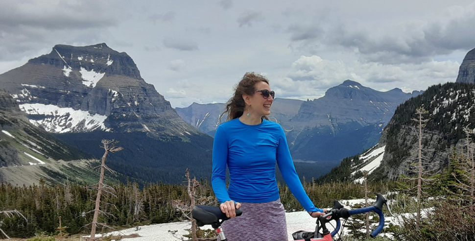 A woman holds a bike and stands in front of large mountains.
