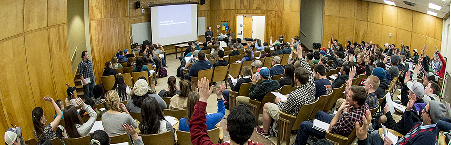Large lecture hall with students raising their hands during a classroom presentation.