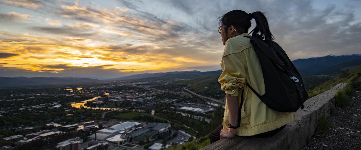 A student with a ponytail sits at the M on Mount Sentinel and looks down on campus and Missoula as a colorful sunset streaks across the sky.
