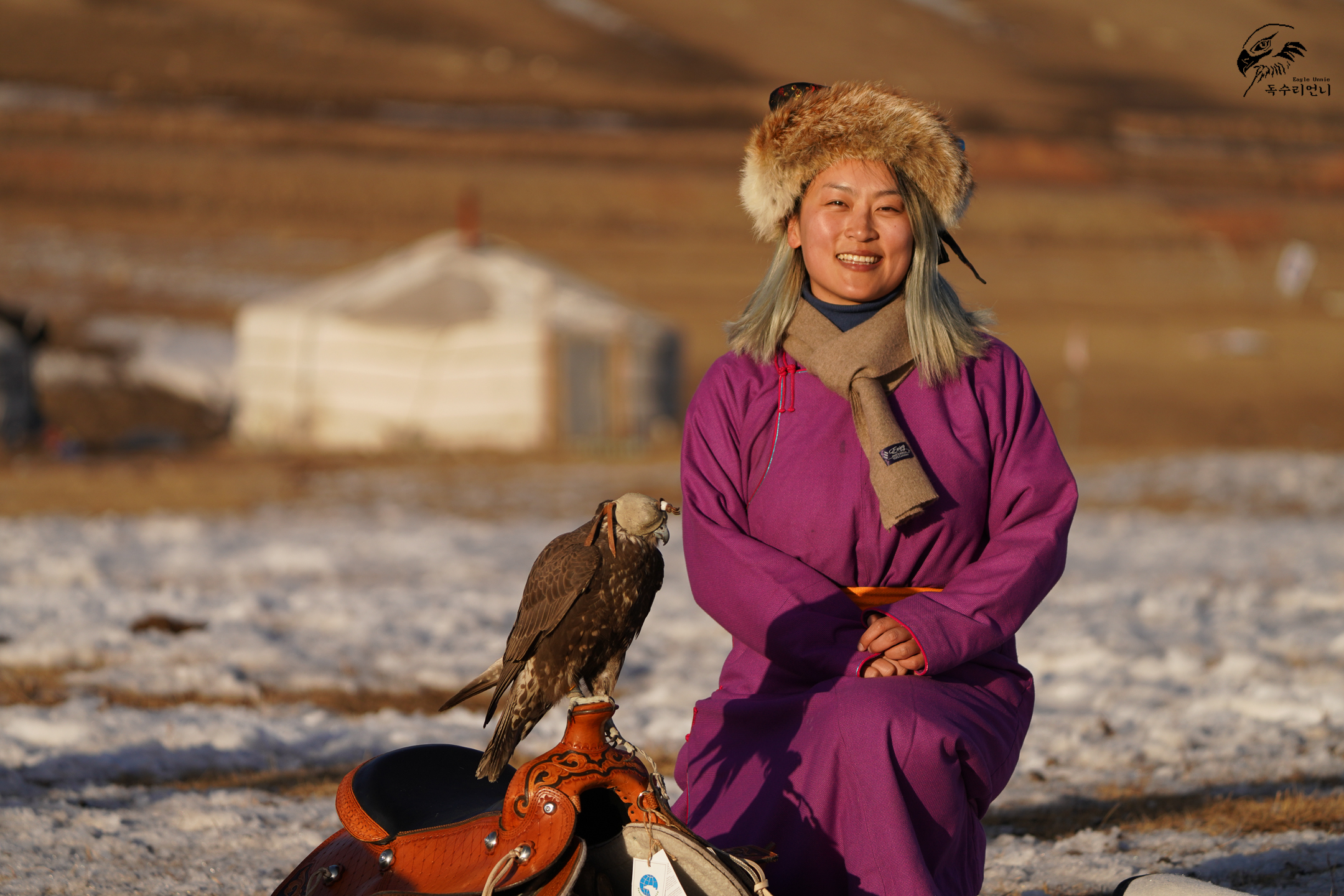 Lee Nari wearing a fur hat and purple coat sitting next to a hawk in a cold field