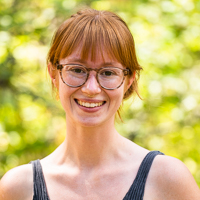 Person smiling in front of green background outside.