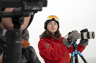 Woman on top of snowy mountain looks to sky with camera in hand.