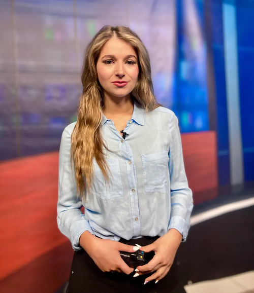 Young woman in button-up shirt stands in front of camera.